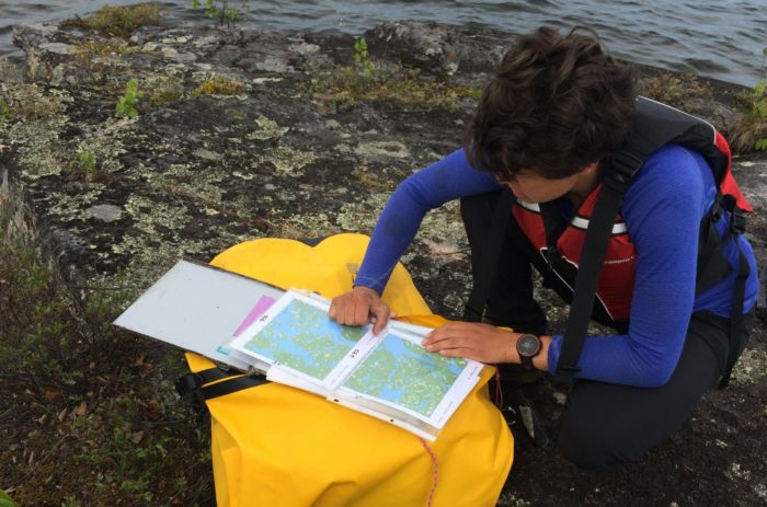 A person wearing a life jacket crouches on the edge of a lake, reading a map